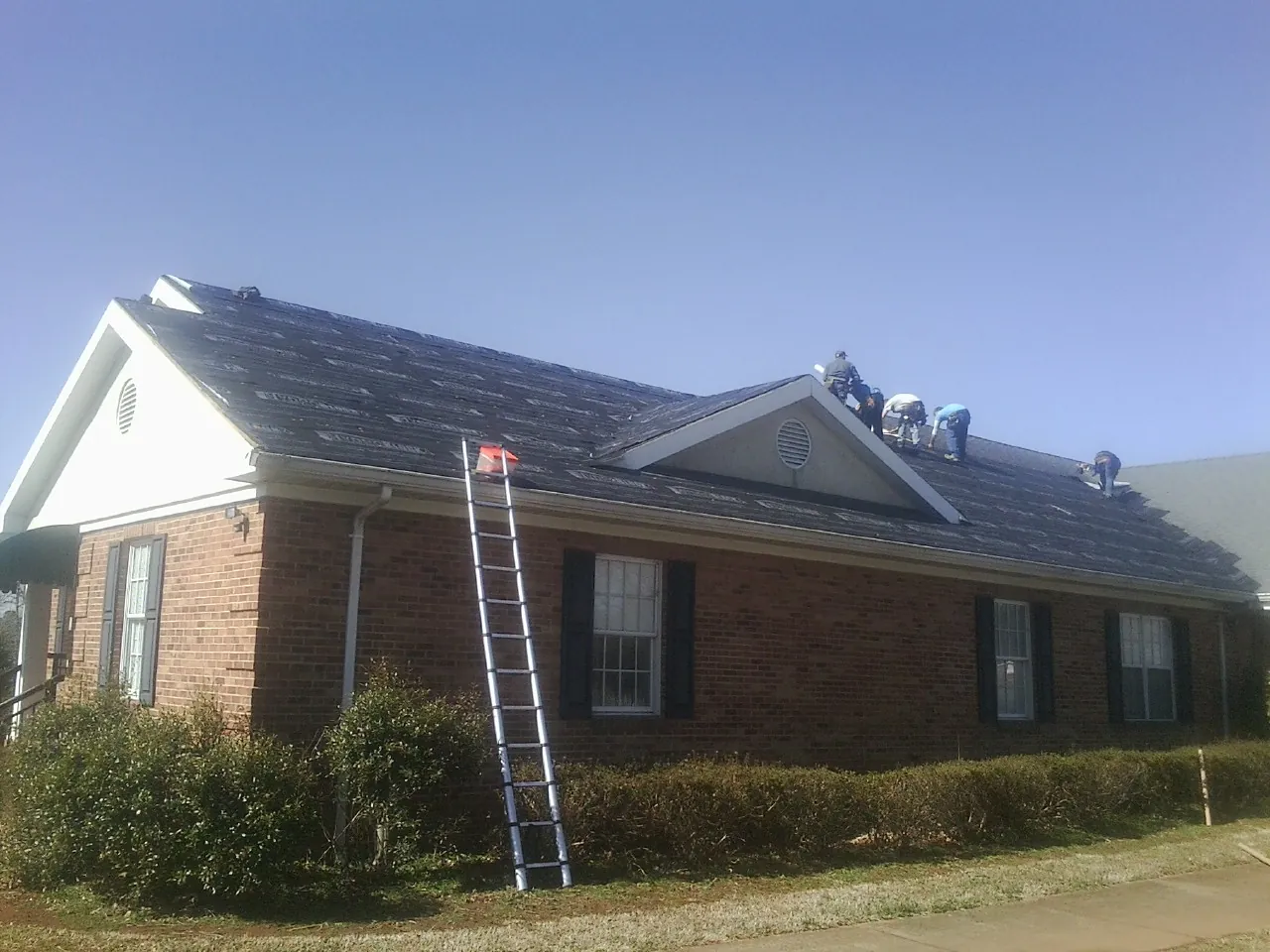 Crew tearing shingles off a brick ranch roof with a ladder set — Metal Roof Repair work by our crew in Horntown