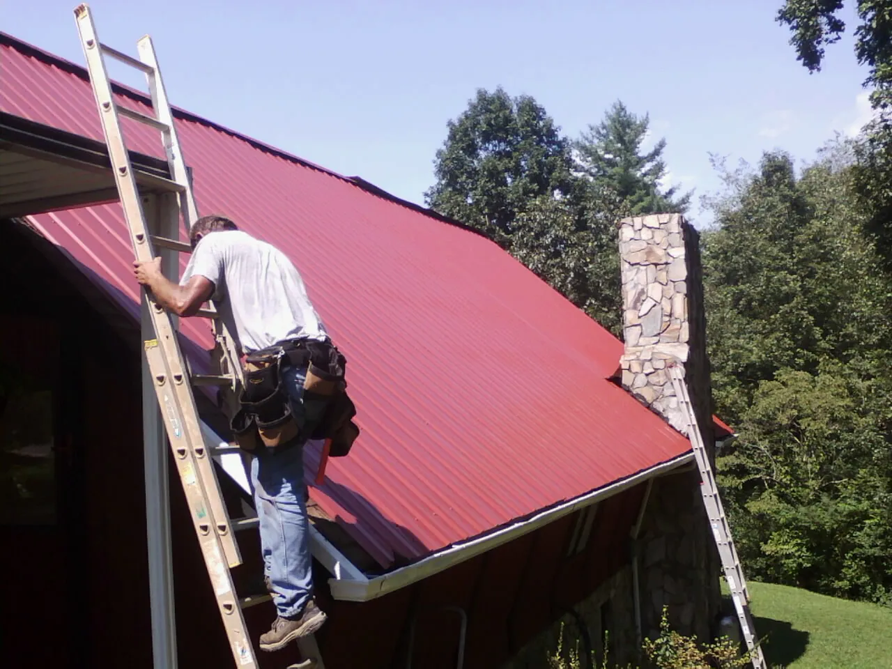 Roofer on a ladder beside a red metal roof and stone chimney — Soffit & Fascia Repair by Roofing Brothers in Horntown